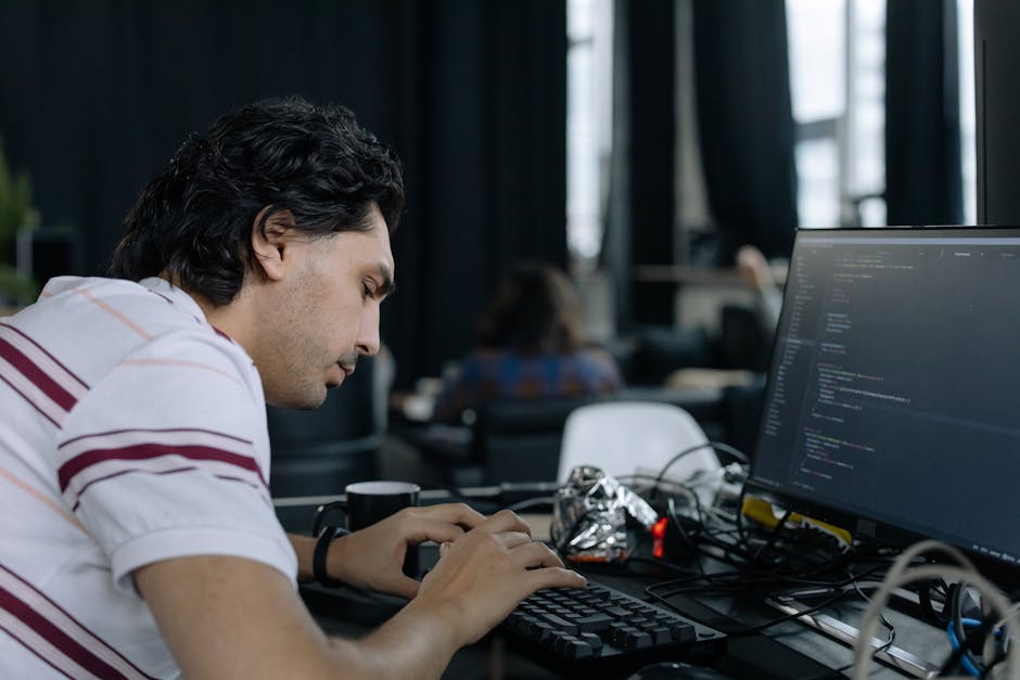Close-up of a person's hands typing on a laptop keyboard, with the screen displaying lines of computer code in a dark-themed text editor. The laptop is positioned on a dark surface, and the background