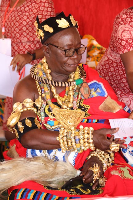 A woman wearing traditional Maasai attire seated outdoors during a cultural event, dressed in vibrant red, blue, and black fabric adorned with intricate beadwork and metal jewelry. She is decorated wi